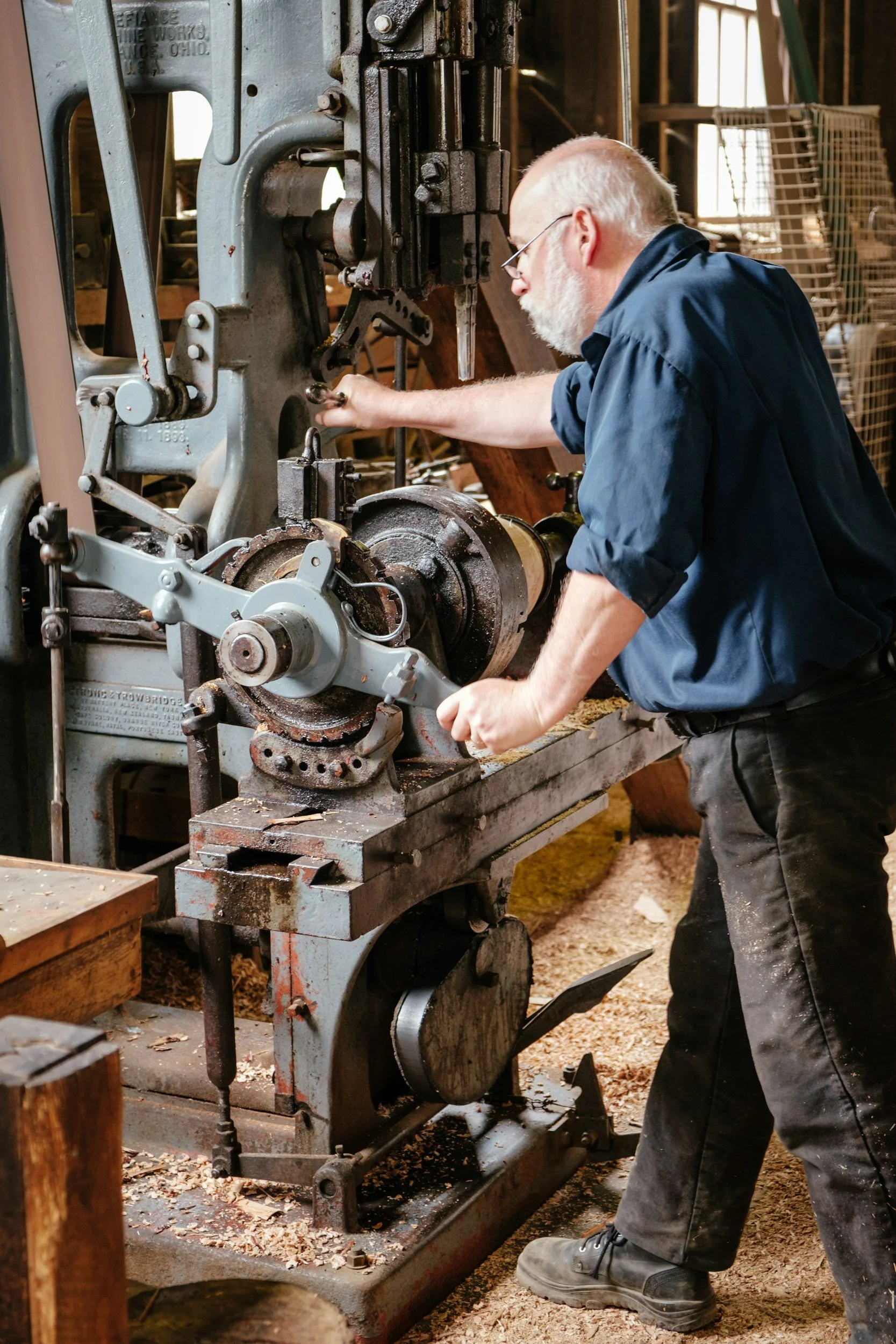 A man with white hair and beard wearing glasses, a dark blue shirt, and black pants operating a vintage band saw in a woodworking shop, surrounded by wood shavings and other woodworking tools.