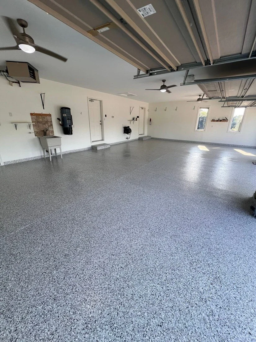 Empty clean two-car garage with garage doors open, ceiling fans, windows, and minimal storage shelves and hooks on the walls.