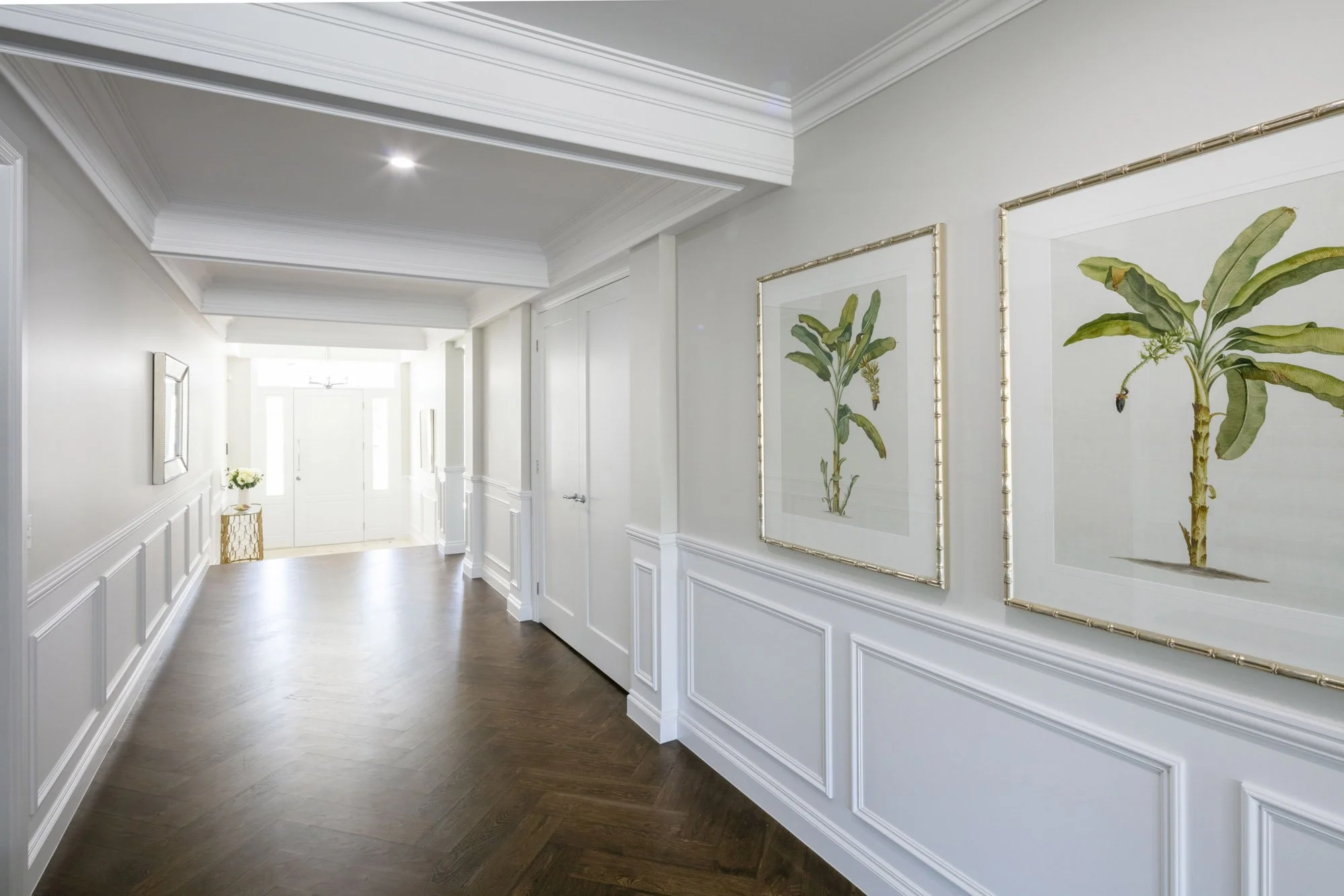 Bright hallway with white walls, crown molding, and framed botanical art prints. Hardwood floors and a small table with a vase of flowers near the door.