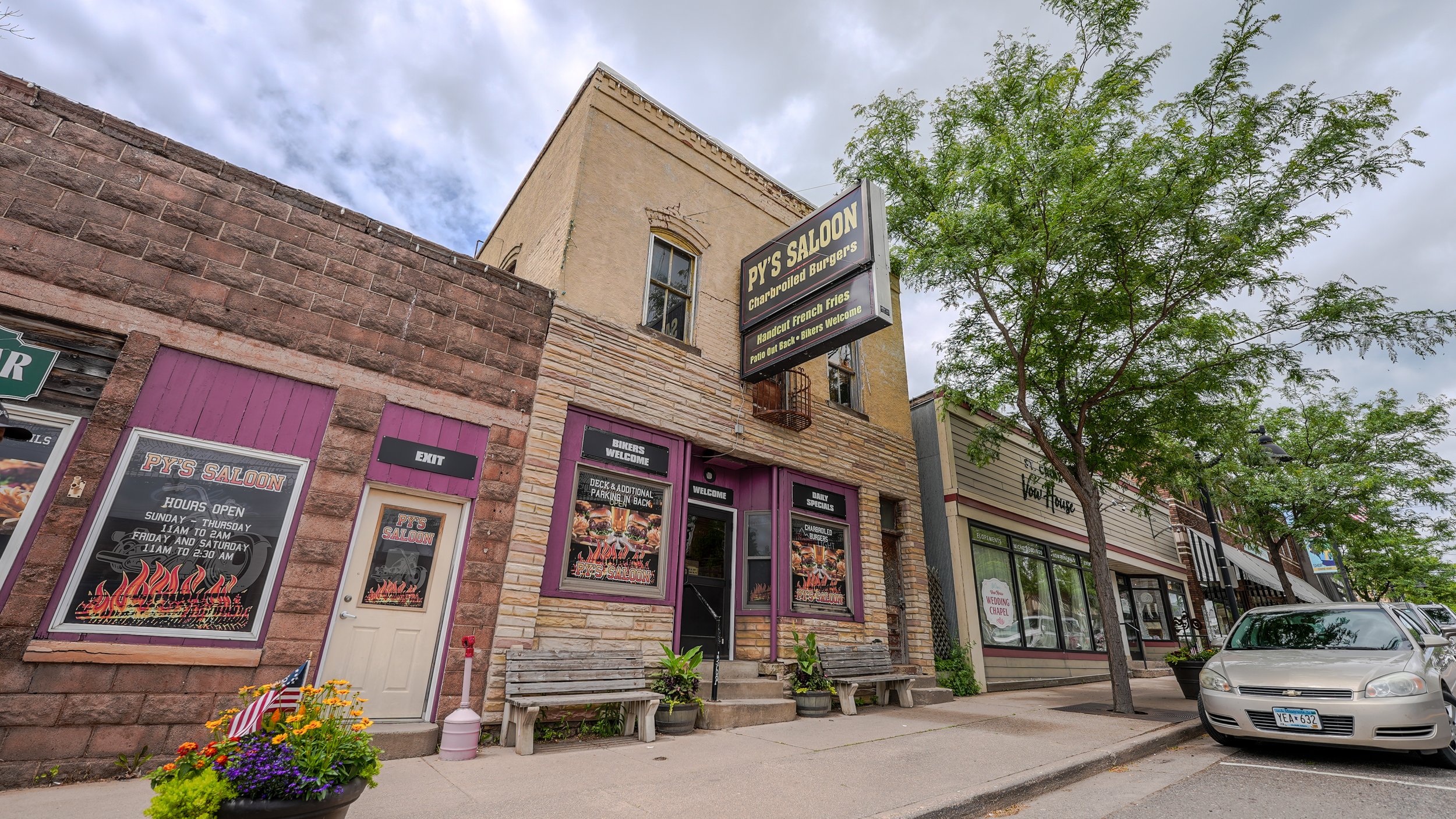 Exterior of Py's Saloon with purple-framed windows and a black and yellow sign, a sidewalk with benches, potted plants, and a tree, under a cloudy sky.