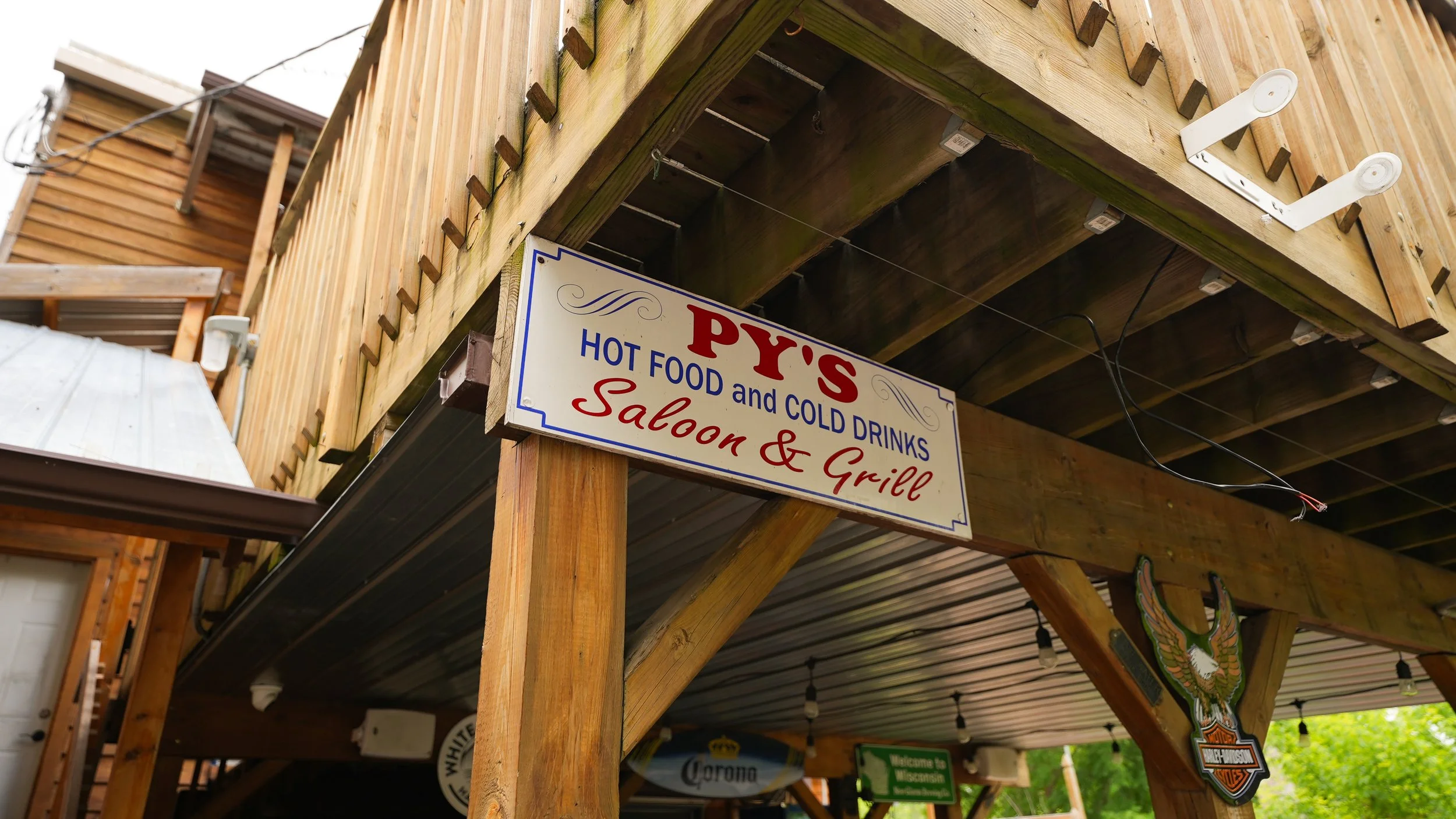 Wooden outdoor bar with a sign that reads "Py's Saloon & Grill," offering hot and cold drinks, with string lights and Harley-Davidson decorations hanging around.