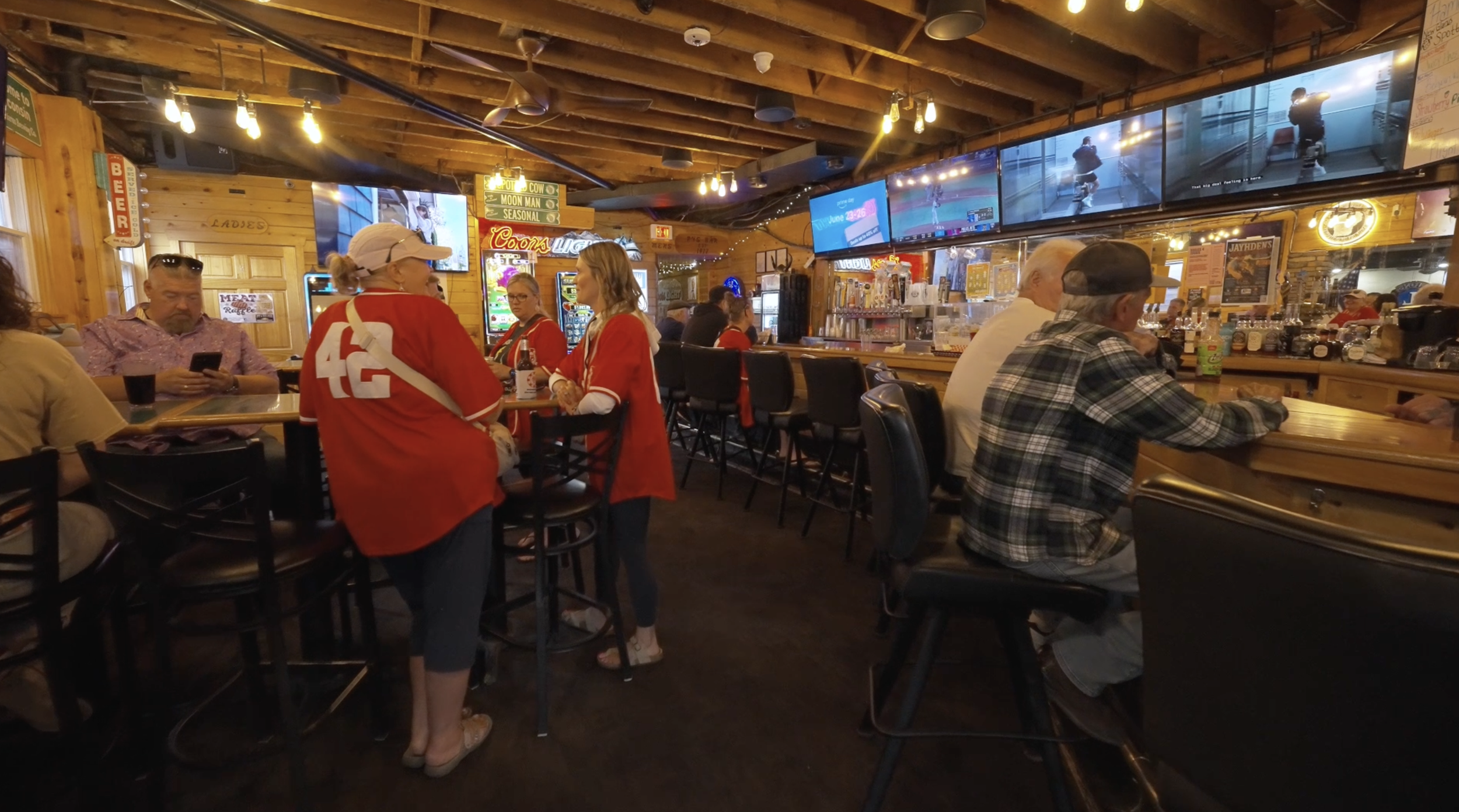 People inside a bar or restaurant with wooden walls and ceilings, watching multiple TVs displaying various sports and entertainment programs, with some patrons seated at a bar and others standing and talking.