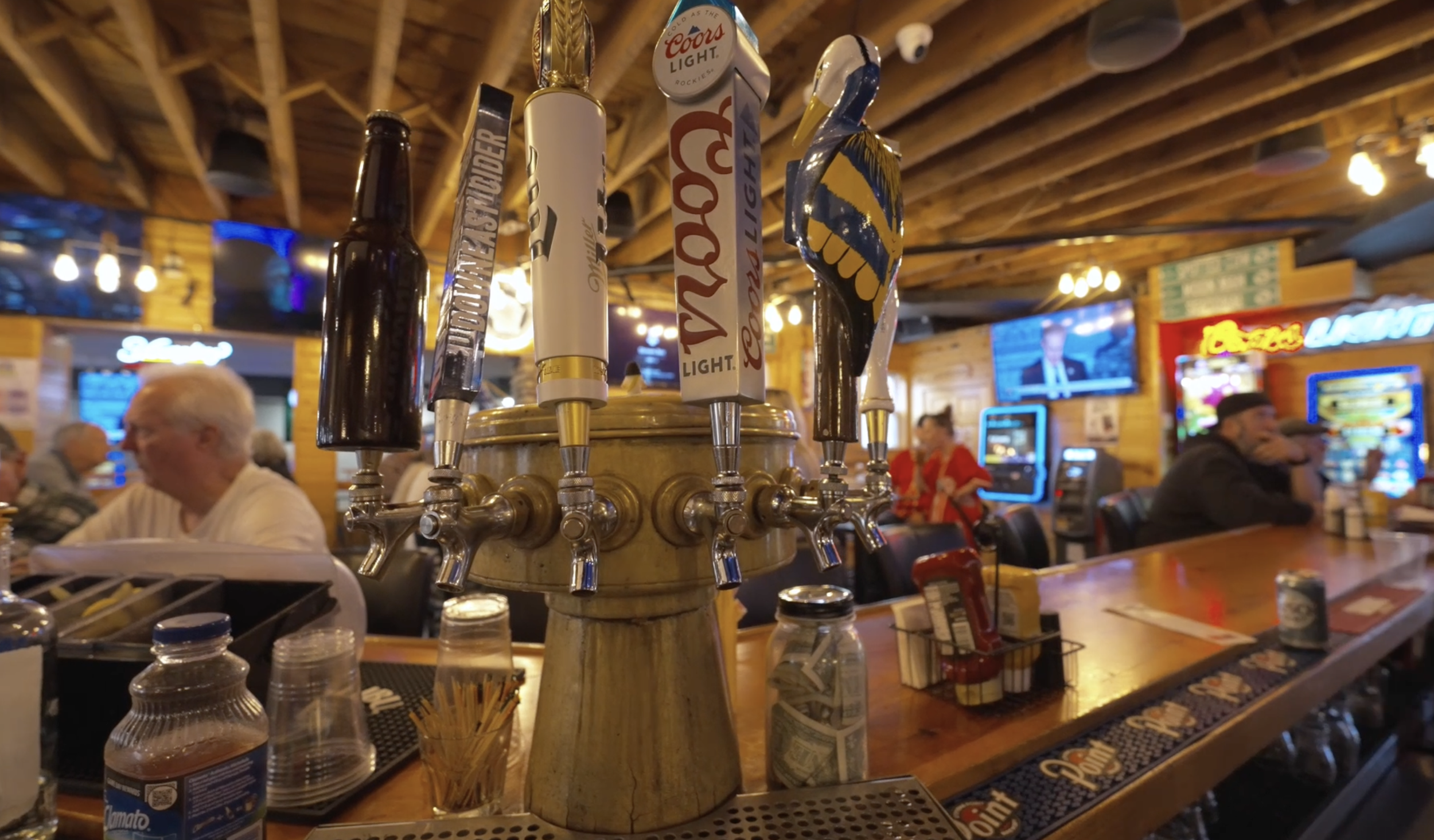 A row of beer tap handles at a bar with a crowded wooden interior and patrons in the background, including an elderly man in a striped shirt and a woman in a red blouse.