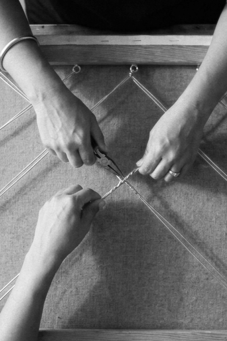 Three hands working together to thread string through a grid of nails on a wooden frame, possibly for a craft or string art project.