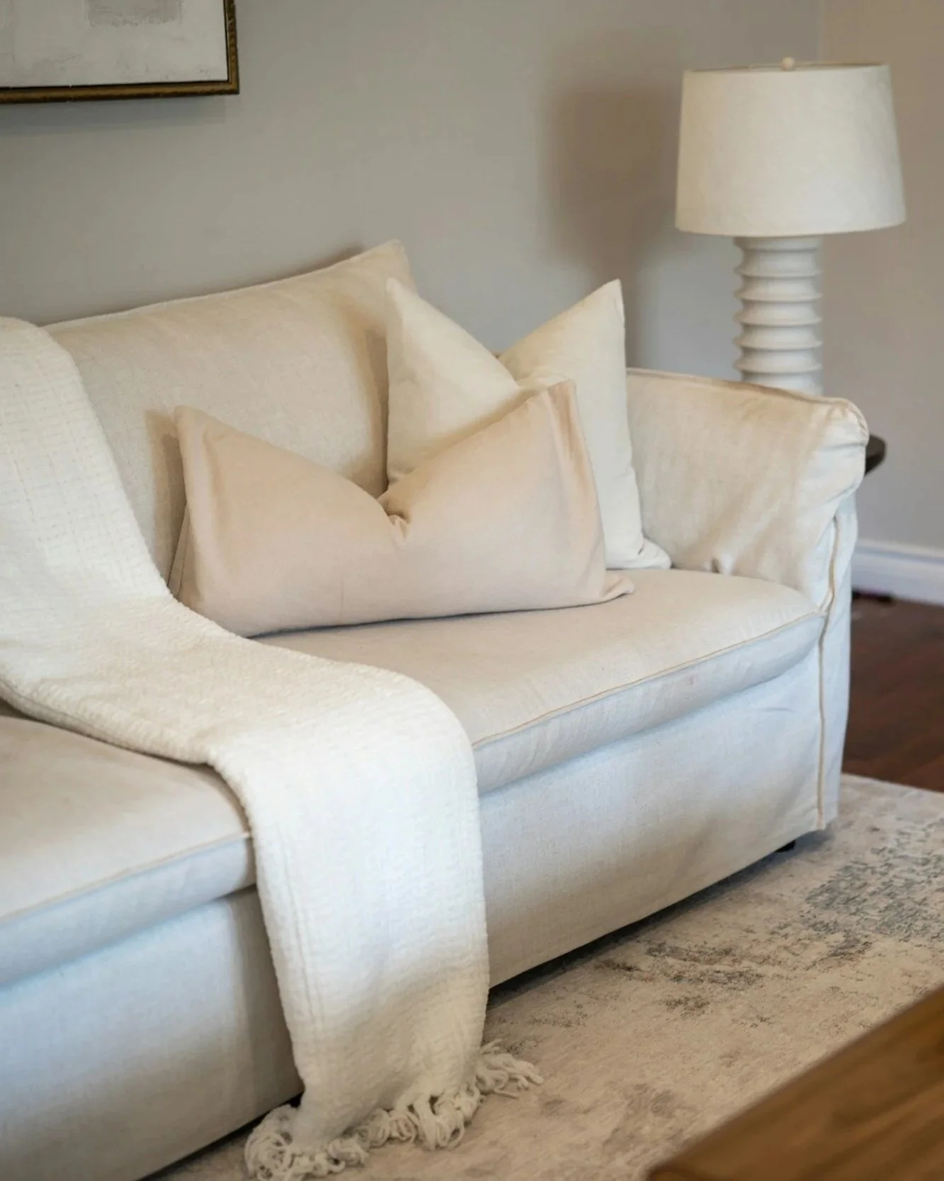 Cream-colored sofa with multiple beige pillows, beige throw blanket on the left armrest, a white textured blanket hanging over the edge, and a beige and white area rug underneath. A white lamp with a rounded, ribbed base is on a side table to the right, with a wall and framed picture partially visible in the background.