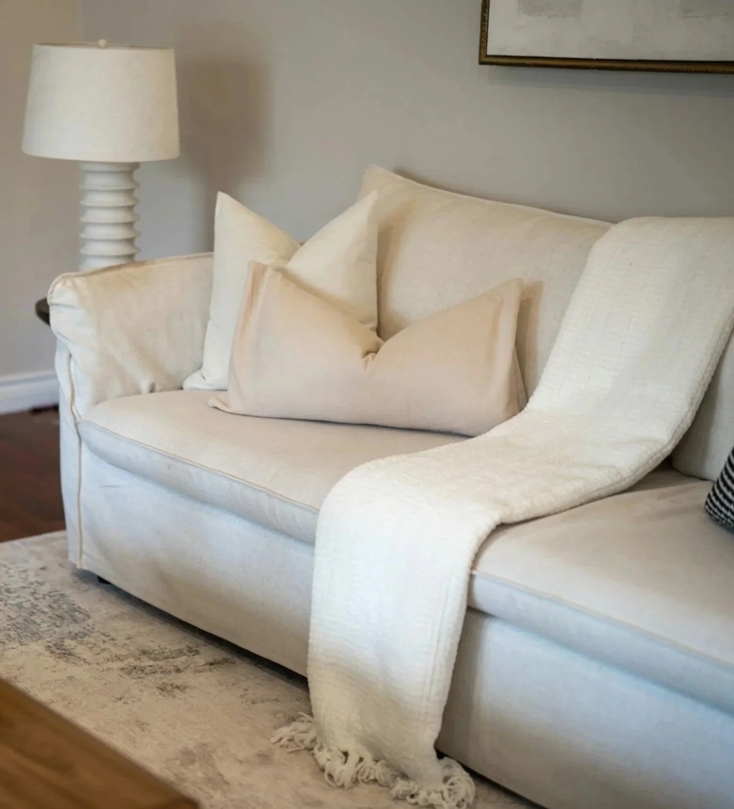 A cream-colored sofa with several matching cushions, a cream throw blanket draped over the arm, a nearby lamp with a white shade, and a rug on a wooden floor in a living room.