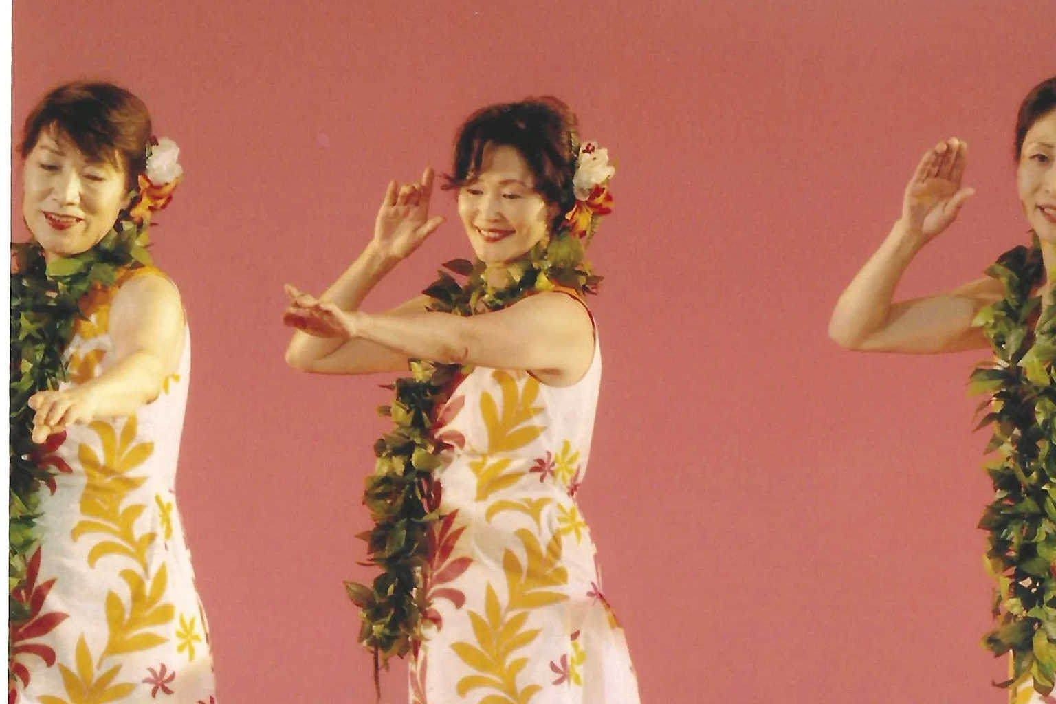 Sakiko, centered, performs hula onstage in Japan with fellow dancers, wearing a white patterned dress, green lei, and flowers in her hair.
