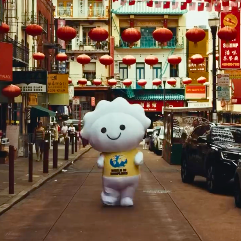Smiling dumpling mascot walks beneath red lanterns along a street in San Francisco’s Chinatown.