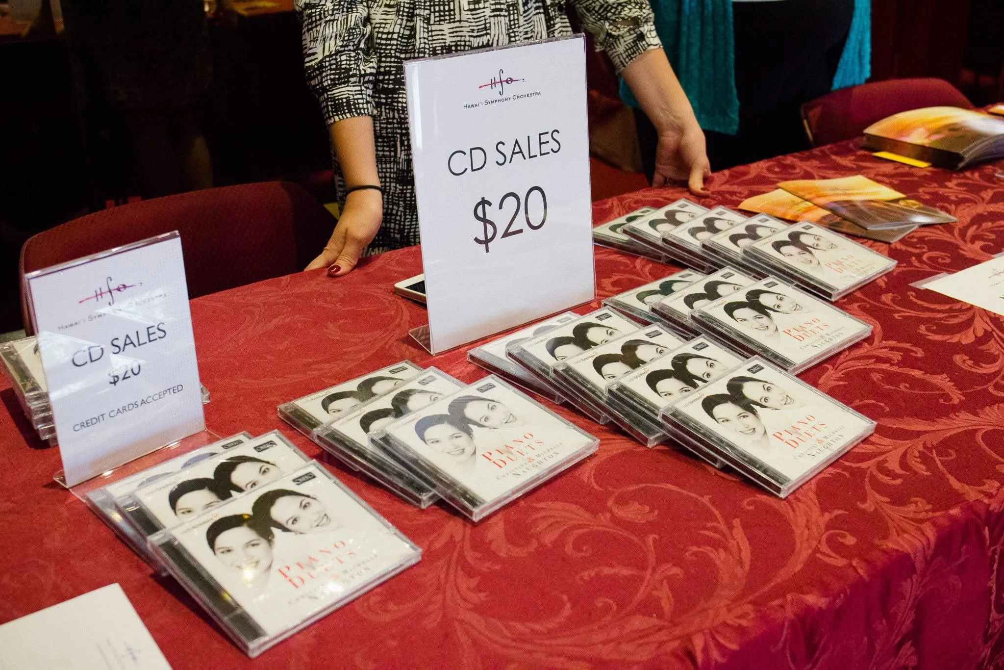Hawaiʻi Symphony Orchestra CD sales table with stacks of recordings, price signs, and a staff member assisting at a concert.