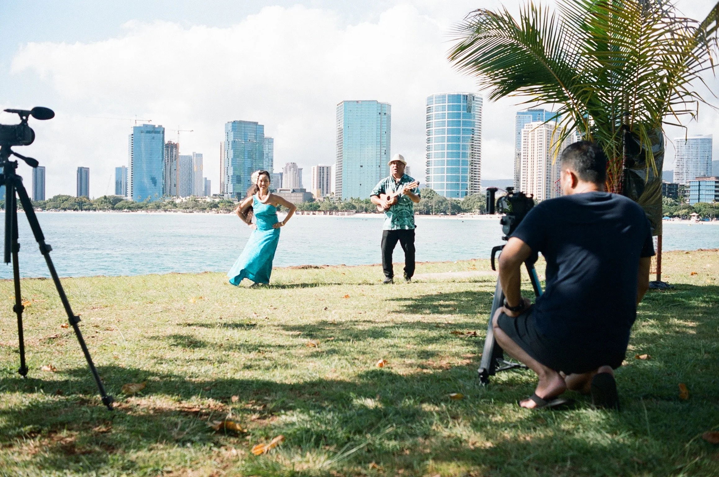 David Pierce’s mother performing hula beside a musician while being filmed at Magic Island on Oʻahu, with the Honolulu skyline in the background.