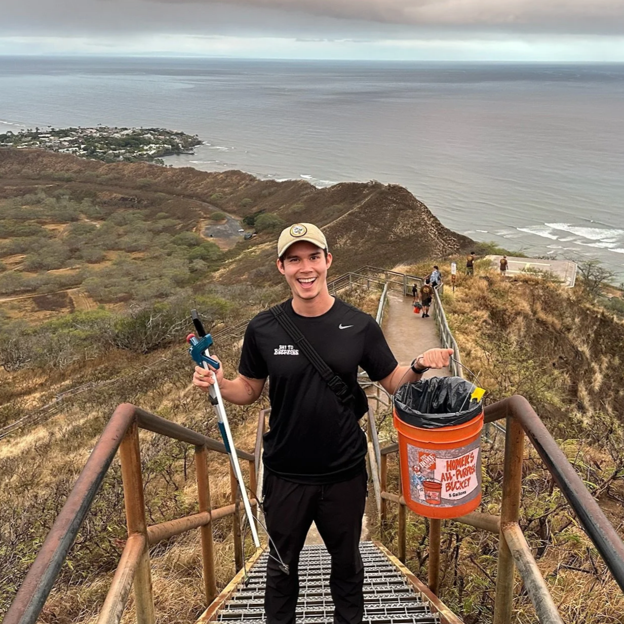 David Pierce collects litter along a coastal trail using a grabber and bucket at Diamond Head State Monument.