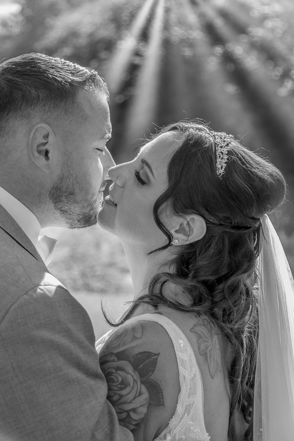 Ein Brautpaar bei einer Hochzeit, in Schwarz-Weiß-Fotografie, nah beieinander stehend, sie schauen sich liebevoll an, im Hintergrund Sonnenstrahlen durch Bäume.