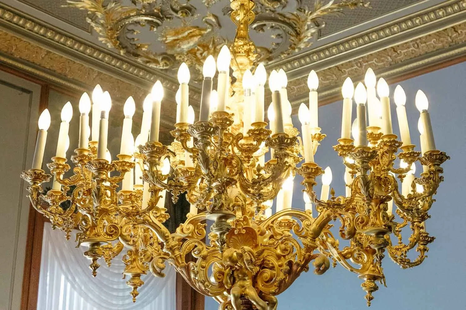 Ein großer, goldener Kronleuchter mit vielen Kerzenleuchten hängt an der Decke eines elegant eingerichteten Raumes.