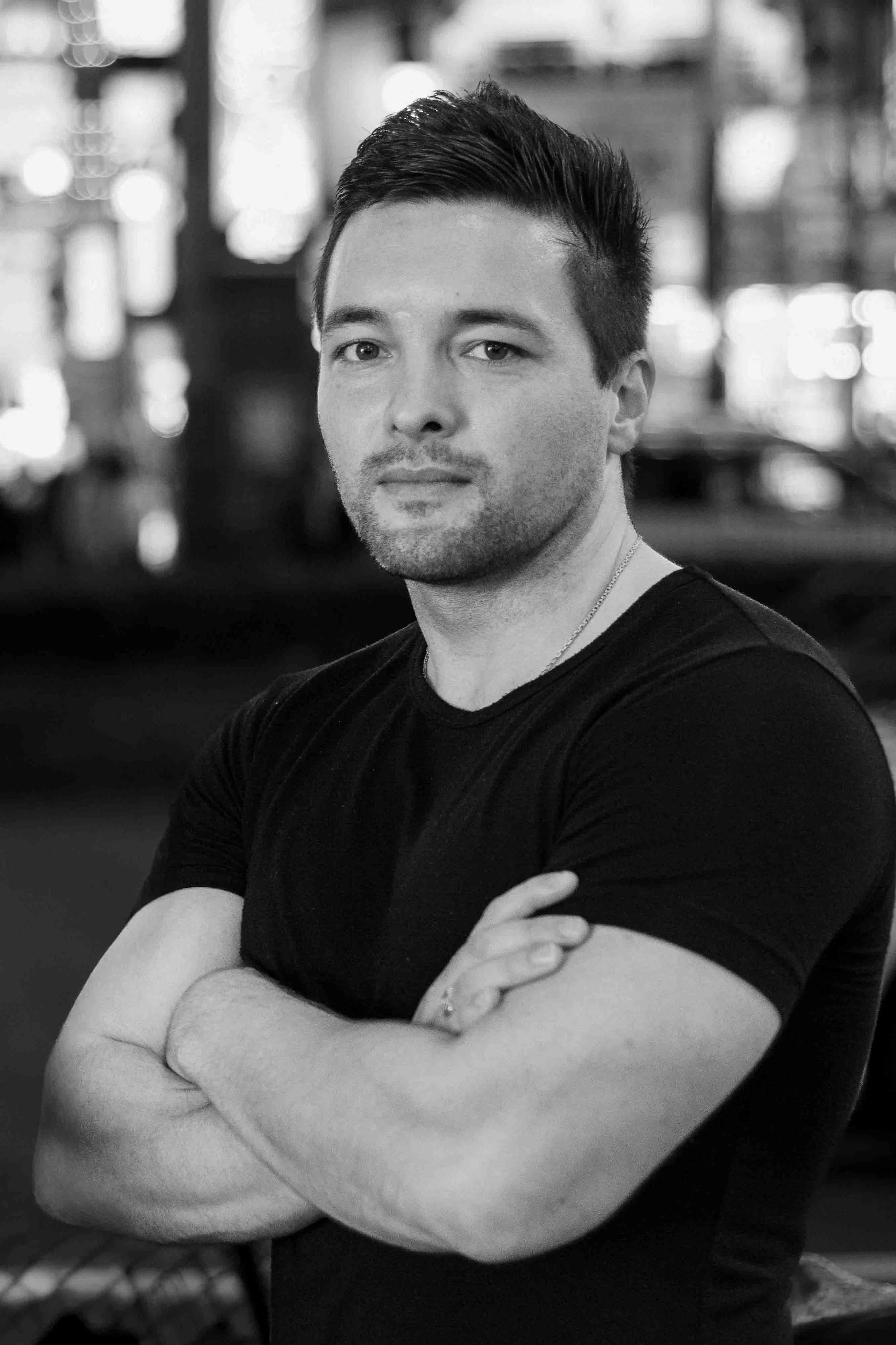 Schwarz-weiß Foto eines Mannes, der die Arme verschränkt trägt, in einem indoor Umfeld, unscharfer Hintergrund.