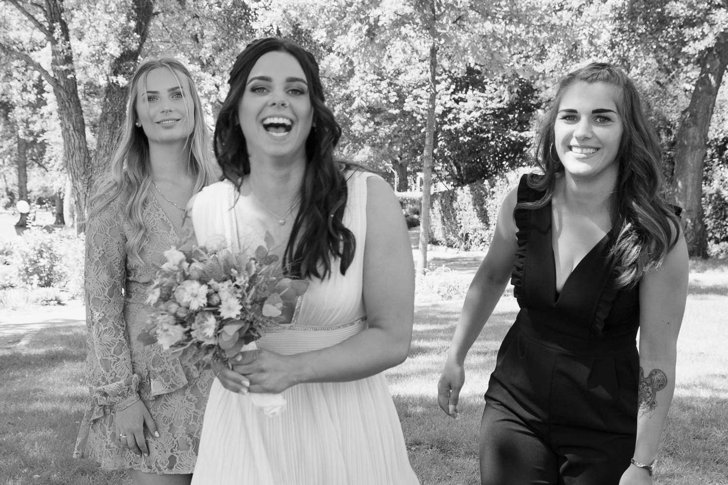 Drei junge Frauen im Park, eine trägt ein Brautkleid, hält einen Blumenstrauß, und die anderen beiden tragen elegante Kleider, alle lacht und haben eine gute Zeit.