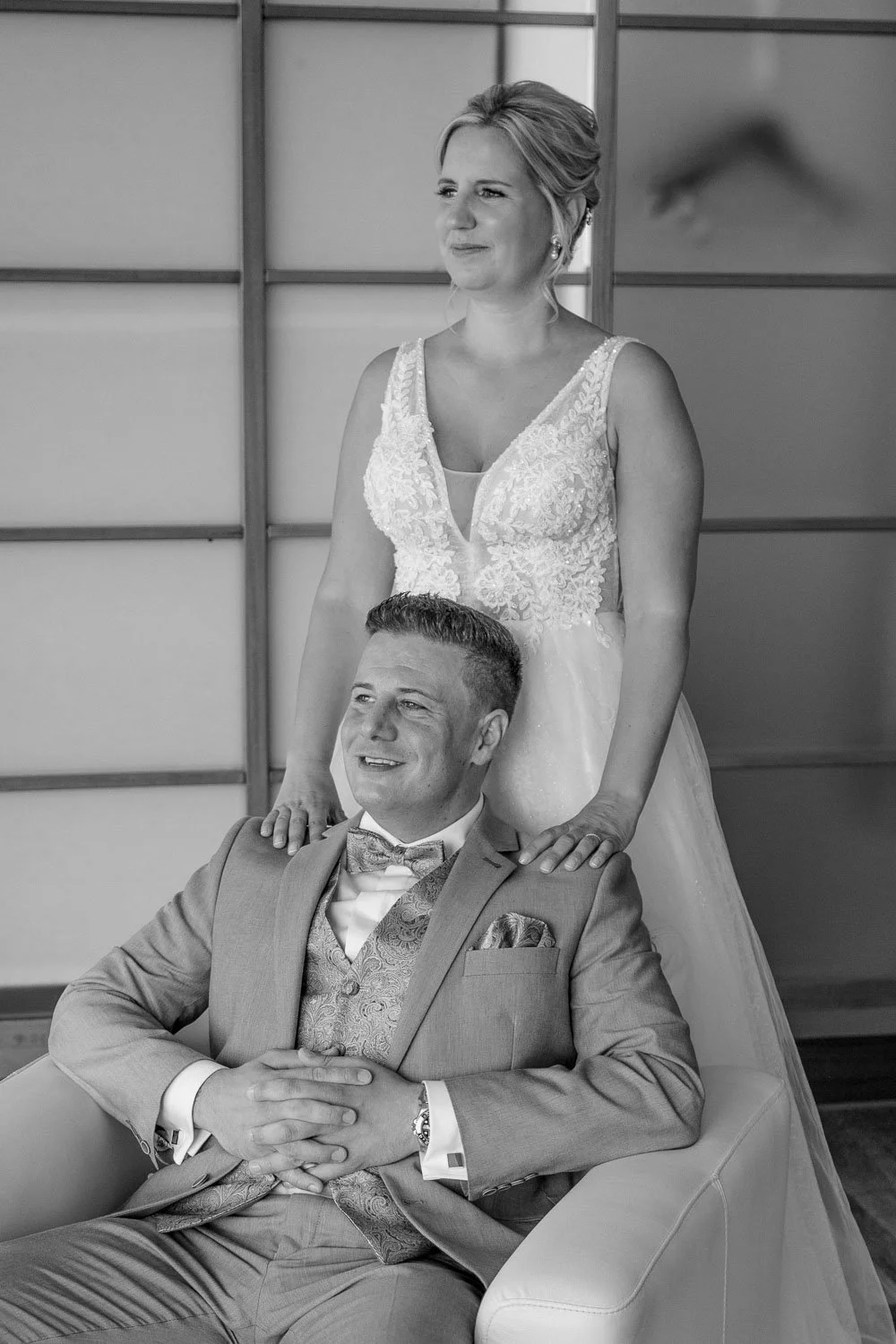 Ein lachendes Paar in formeller Kleidung bei einer Hochzeit, die Frau steht hinter dem Mann, der auf einem Sofa sitzt, beide vor einem modernen Raum mit Holz-Wandpaneelen.
