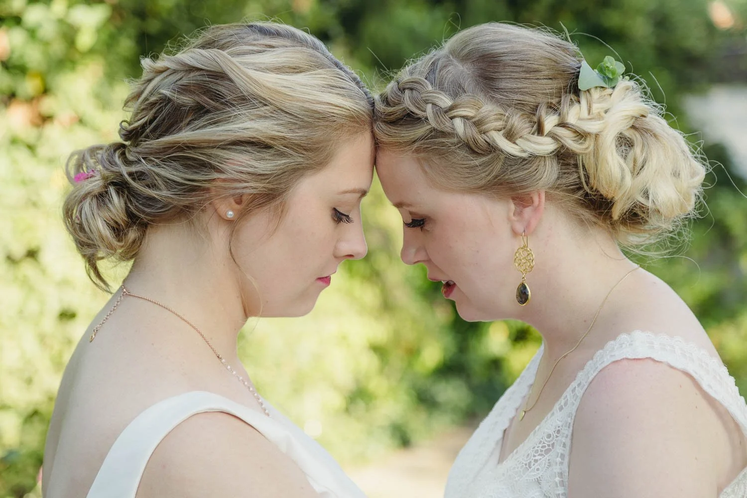 Zwei Frauen mit verschränkten Stirnen, die sich berühren, bei einem Außenauftritt, mit Brautkleidern und lockigem, blondem Haar, umgeben von Grün.