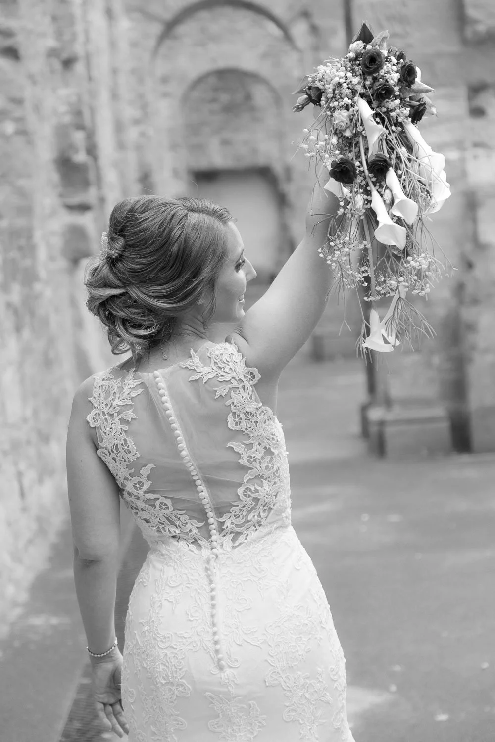 Schwester oder Braut hält an einem Hochzeitstag einen Blumenstrauß in der Hand, trägt ein elegantes, detailliertes Hochzeitskleid, im Hintergrund eine alte Steinmauer.