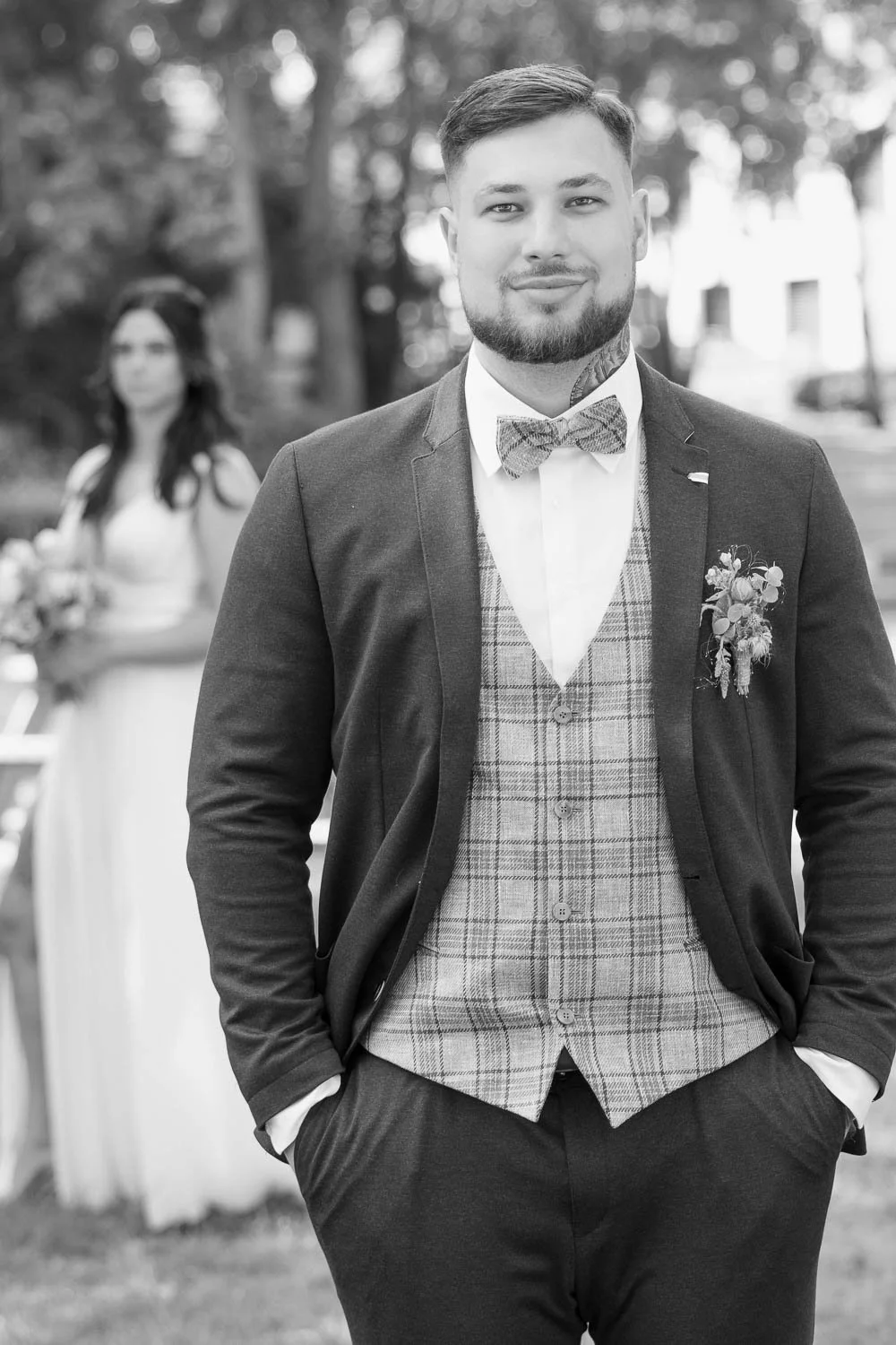 Schwarz-weiß-Foto eines Mannes in formeller Kleidung mit Anzug, Weste, Krawatte und Blume am Revers, im Hintergrund eine Frau in Hochzeitskleid und Brautstrauß bei einer Hochzeit im Freien.