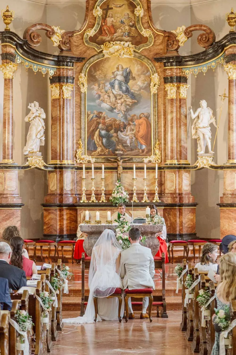 Hochzeitszeremonie in einer Kirche mit Braut und Bräutigam, die vor einem Altar sitzen. Die Kirche ist reich verziert mit Statuen, Kerzen und religiösen Gemälden an der Wand hinter dem Altar.