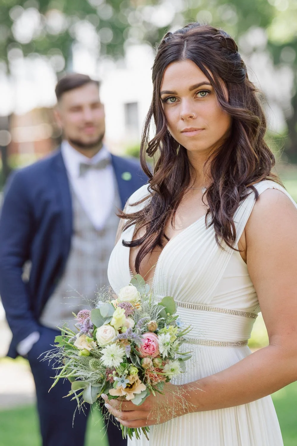 Junge Frau in Hochzeitskleid mit Blumenstrauß vor einem Mann in Anzug bei einer Hochzeit im Freien.