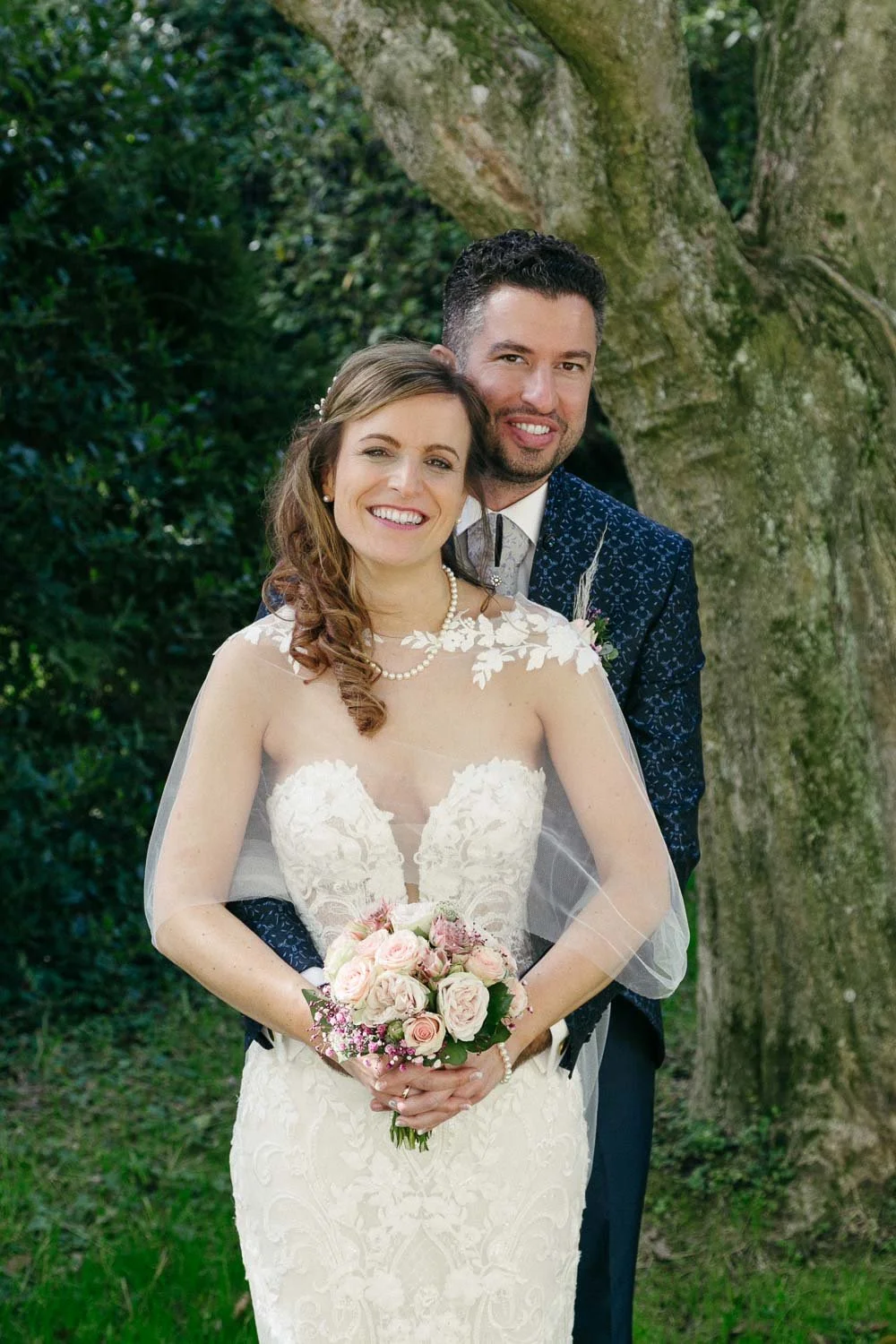 Ein glückliches Brautpaar vor einem Baum bei einer Hochzeit, die Frau trägt ein weißes Spitzenkleid und hält einen Blumenstrauß, der Mann trägt einen dunkelblauen Anzug.