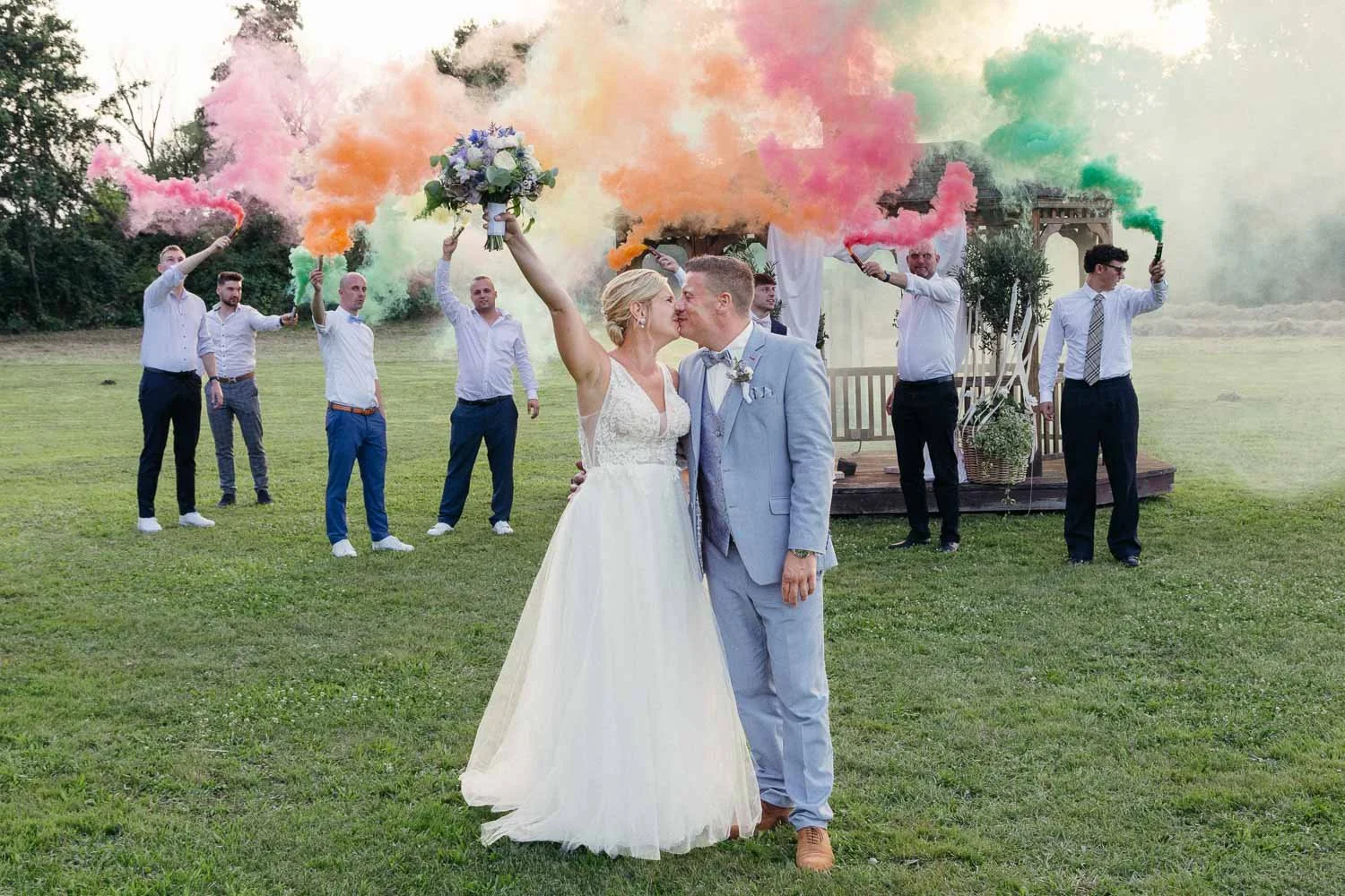 Ein Brautpaar küsst sich, während im Hintergrund Rauchfackeln in bunten Farben gehalten werden, bei einer Hochzeit im Freien.