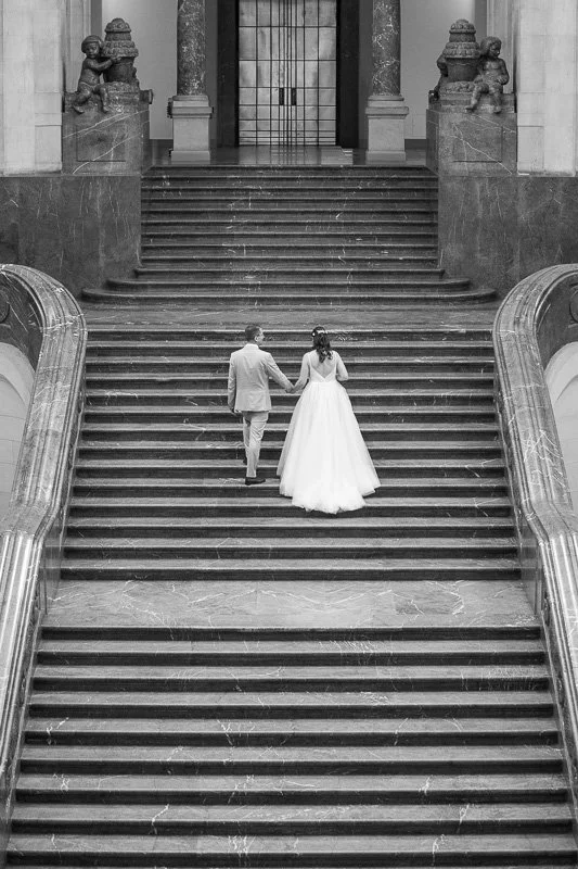 Ein Brautpaar hält sich an Hand auf einer großen Treppe in einem eleganten Gebäude, mit schwarzen Steinstufen und barocken Verzierungen.