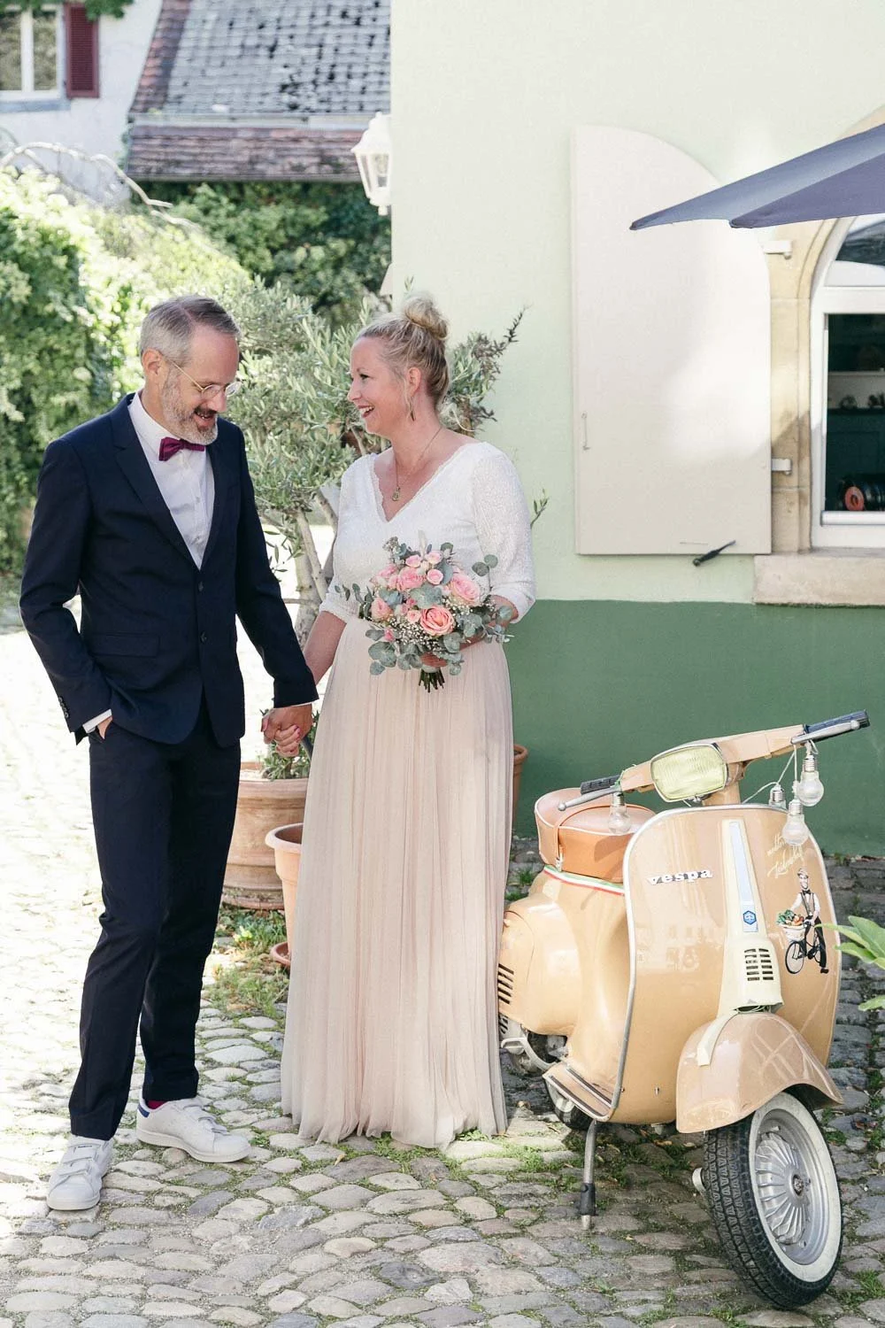 Ein verliebtes Paar an einem Hochzeitstag steht auf einem Kopfsteinpflaster vor einer grünen Hauswand. Sie hält eine Blumenstrauß, er trägt einen Anzug mit Fliege. Es ist sonnig, und im Hintergrund sind Bäume und das Dach eines Hauses zu sehen, neben