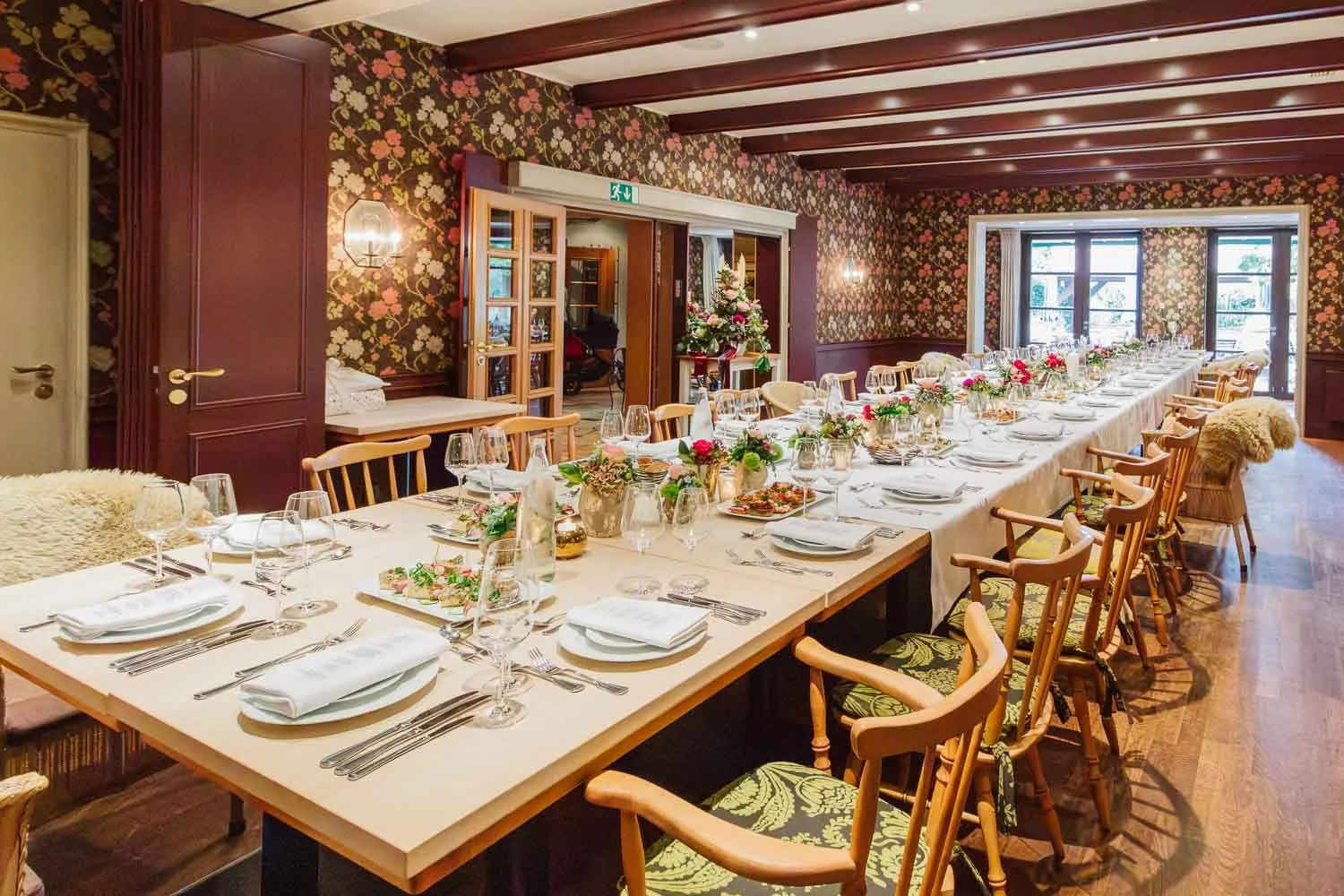 Ein festlich gedeckter langer Esstisch in einem stilvollen Raum mit floral gemusterten Wänden, Blumenarrangements und Geschirr für ein Dinner-Ereignis.