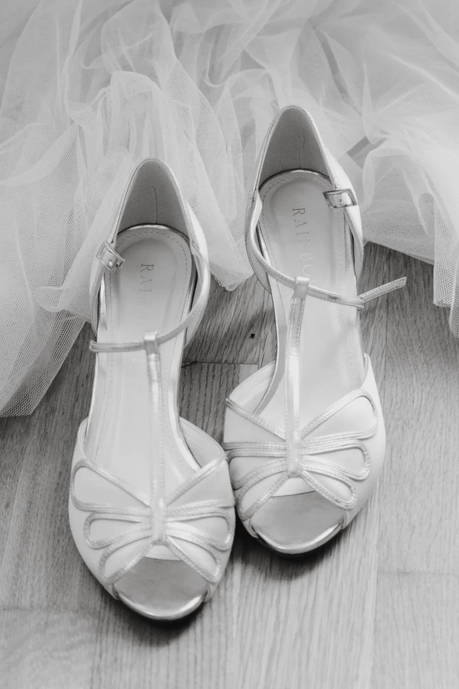Weiße High Heels mit Schleifen, auf Holzfußboden, daneben weiße Tüllstoff.