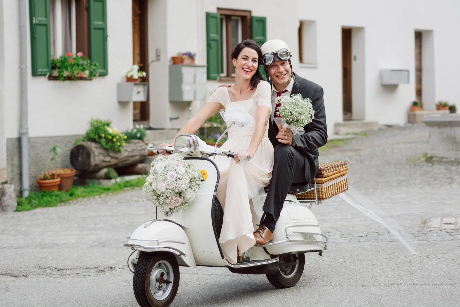 Ein Brautpaar fährt auf einem weißen Vespa-Roller durch eine ruhige Straße. Die Braut trägt ein weißes Kleid und hält einen Blumenstrauß, während der Bräutigam in einem schwarzen Anzug sitzt, einen Helm trägt und ebenfalls einen Blumenstrauß hält. Die Vespa ist mit Blumen dekoriert, und im Hintergrund sind Häuser mit grünen Fensterläden und Blumen zu sehen.