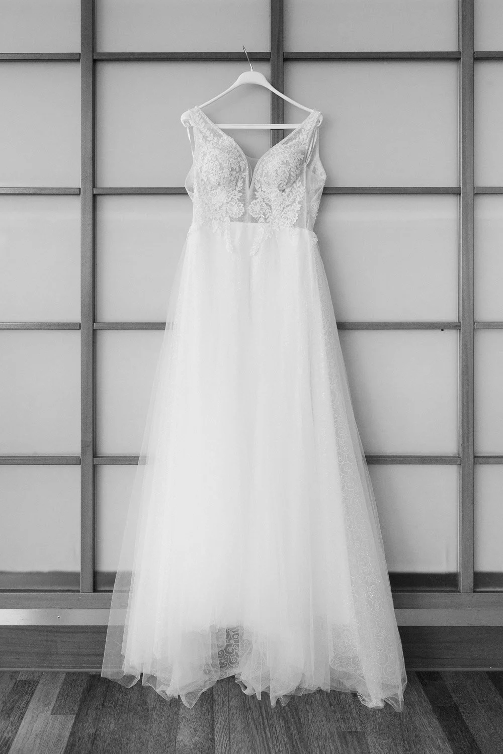 Weiße Brautkleid mit Spitzen-Details hängt an einem weißen Kleiderbügel vor einer grauen Holzpaneele.