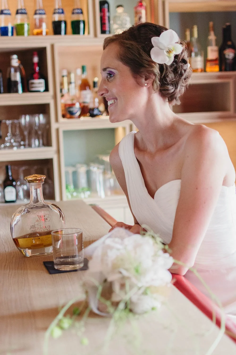 Eine lachende Frau mit braunen, lockigen Haaren und Orchideenblume im Haar sitzt an einer Bar, im Hintergrund sind Alkoholflaschen und Gläser sichtbar.