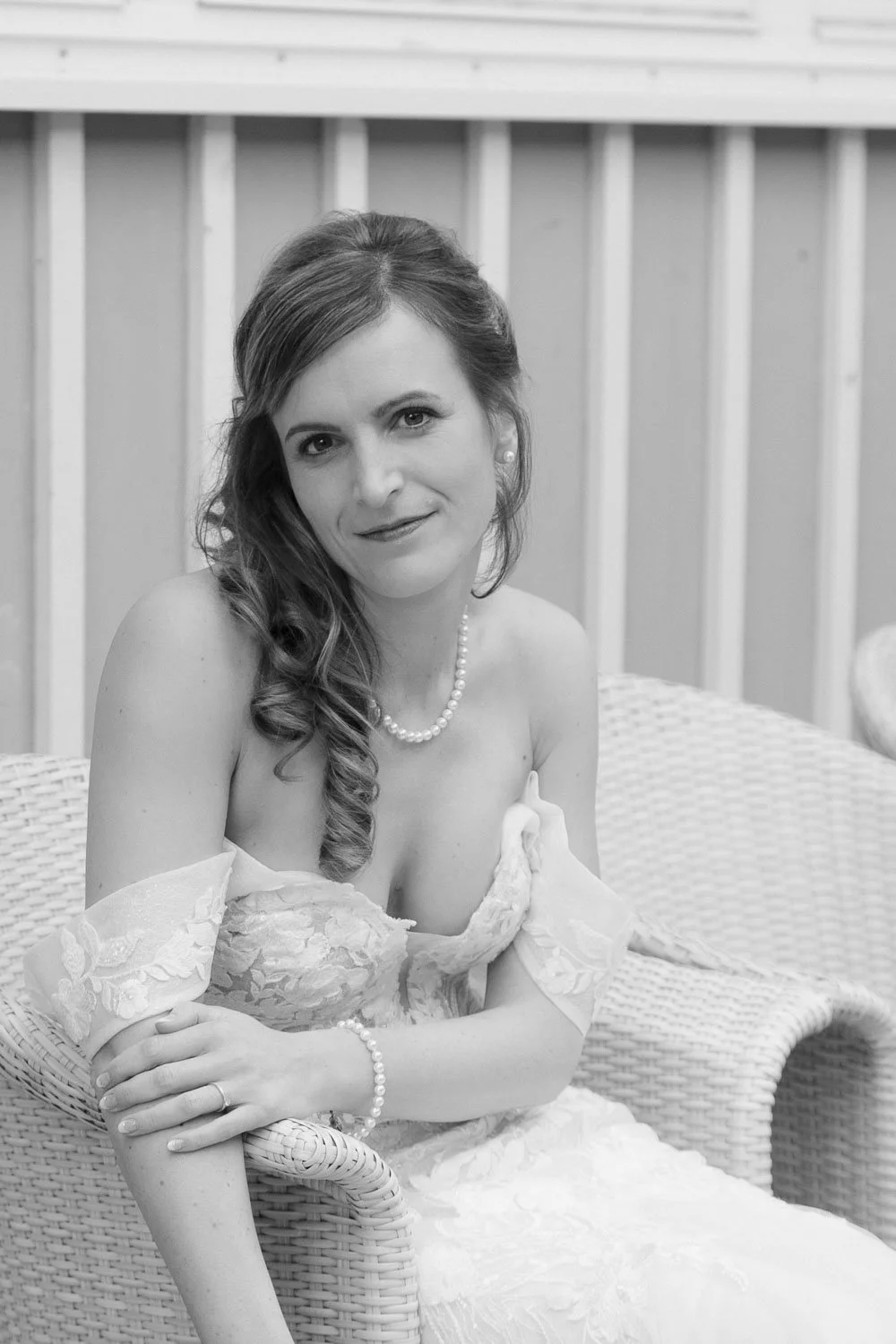 Schwarz-weiß-Foto einer Frau in eleganter Hochzeitstracht, sitzend auf einem Korbsessel, mit Lockenfrisur, Perlenkette und Armband, vor einem Holzpaneel-Hintergrund.
