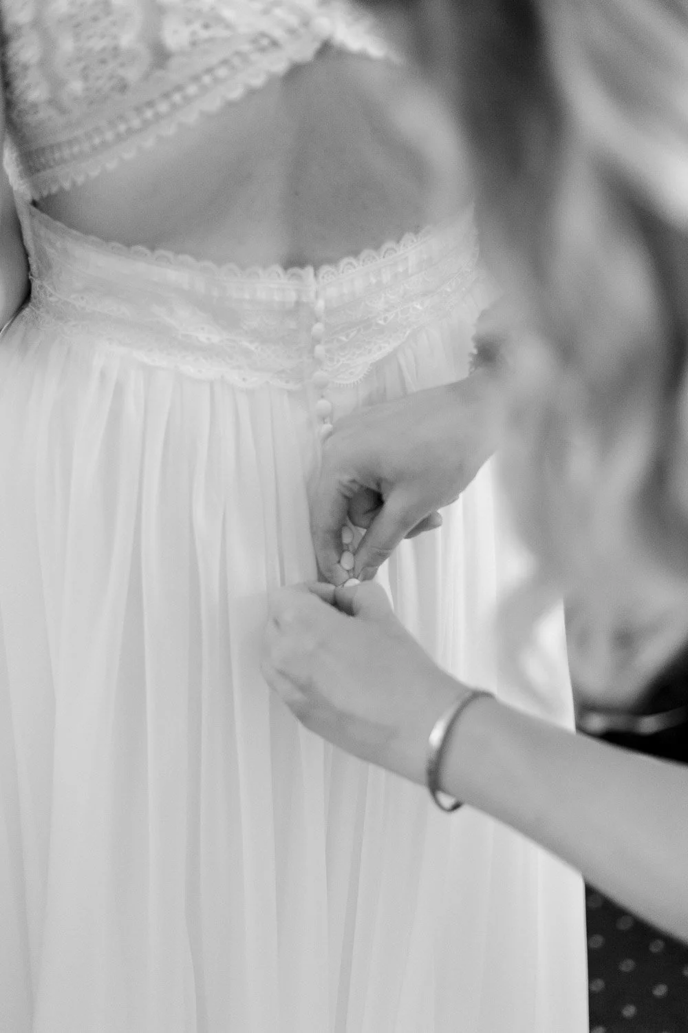 Eine Frau zieht bei einer Braut das Hochzeitskleid zu, das mit Spitze und Knöpfen verziert ist.
