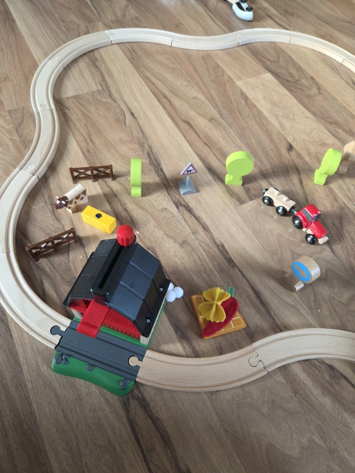 Spielzeuggleis mit Bauernhofthema, Holzschienen, Tiere, Bäume, Tiere, eine kleine Lokomotive und Fahrzeuge auf einem Holzboden.