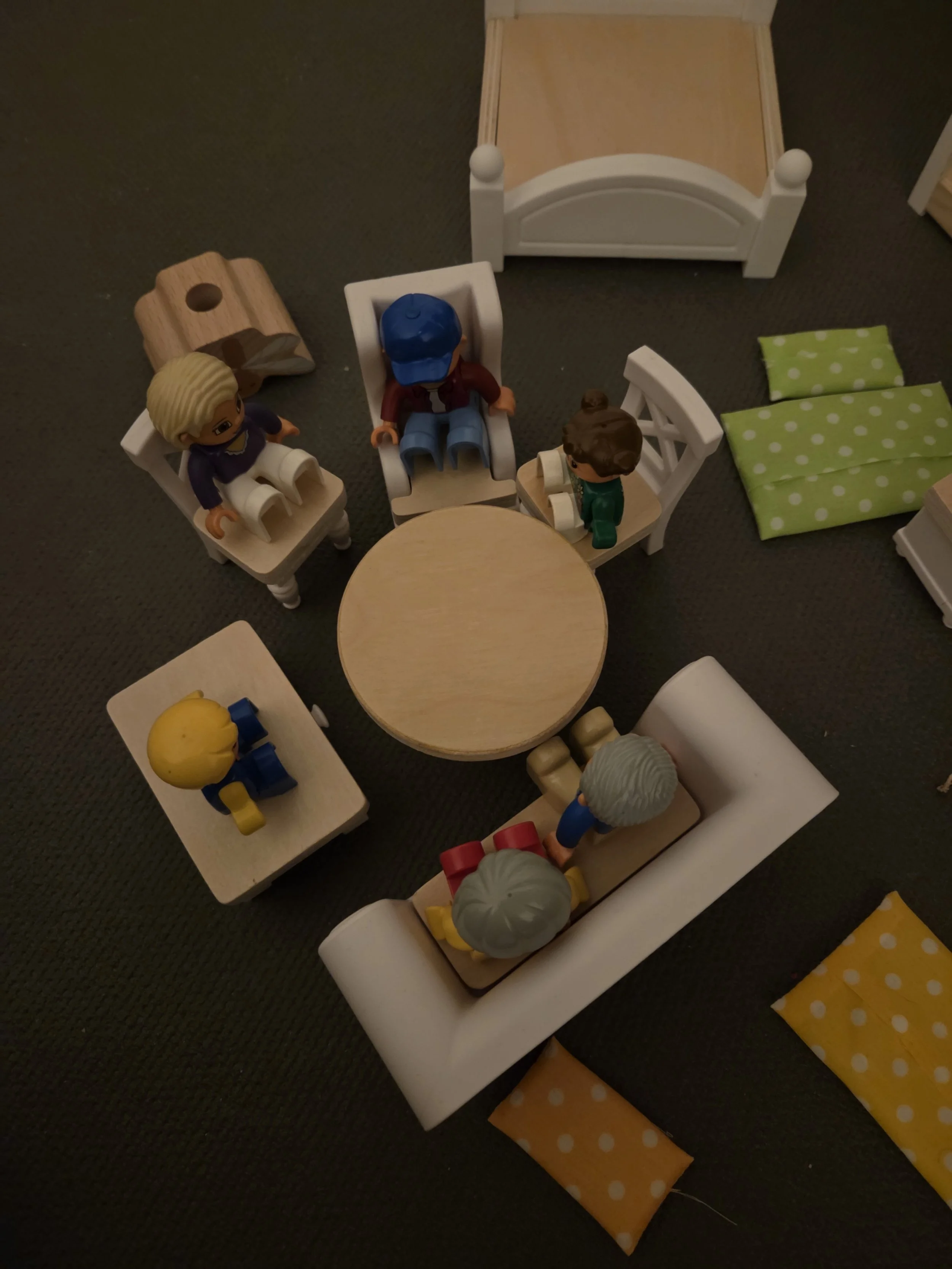 Miniaturfiguren sitzen auf kleinen Stühlen und einer Bank um einen runden Tisch in einer Puppenecke. Es gibt auch eine Mini-Puppenbank und Polster mit grünen und gelben Polka-Dot-Mustern.