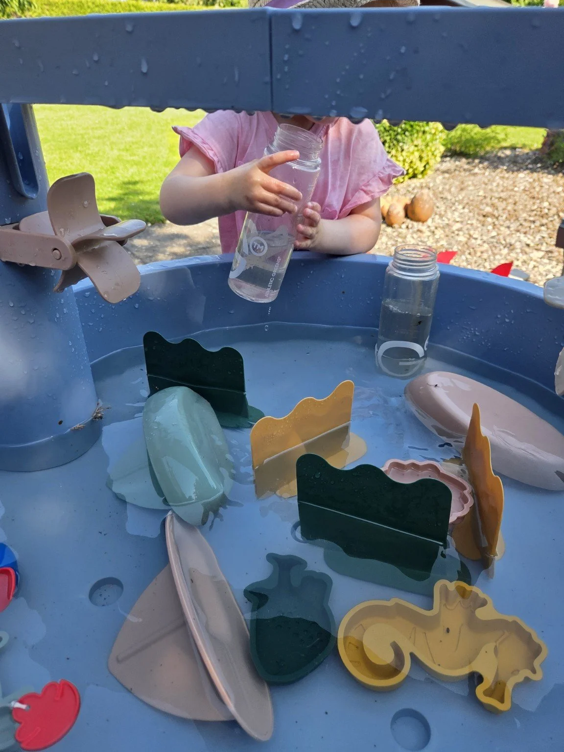 Ein Kind spielt mit Wasser und bunten Seifenblasenformen auf einem blauen Wasser- und Seifenblasentisch im Garten.
