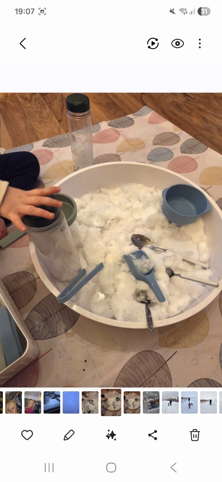 Ein Kinderhand benutzt einen Löffel, um Eis aus einem großen, runden Behälter zu holen, um es zu essen. Es sind auch mehrere Silikonlöffel und ein Trinkbehälter auf einer bunten Tischdecke mit Mustern. Im Hintergrund ist ein Holzboden.