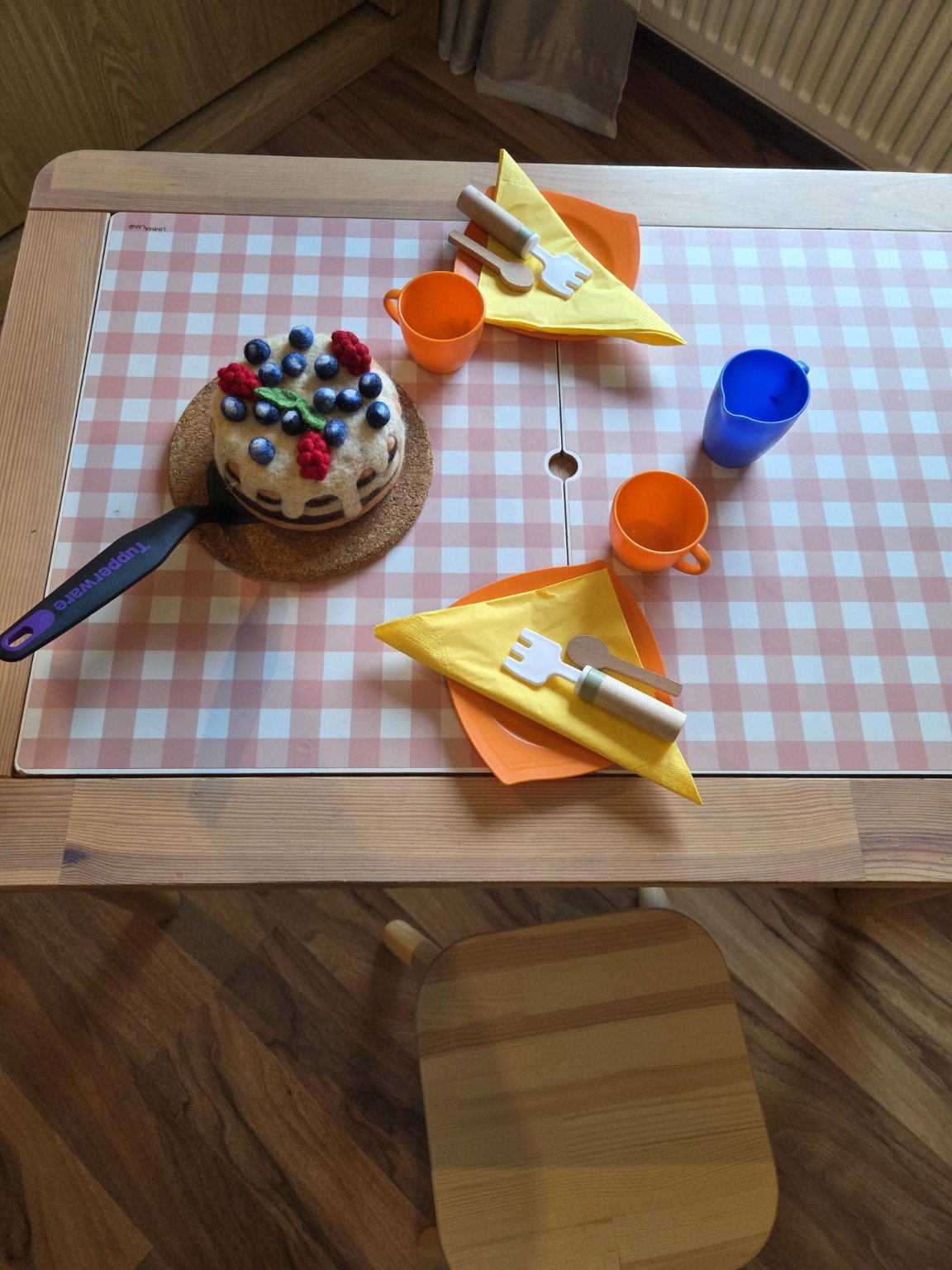 Ein Kindertisch mit einer Mini-Torte, Papptellern, -bechern, Gabeln, Messern, und Servietten; die Torte ist mit Beeren und einer grünen Dekoration verziert.