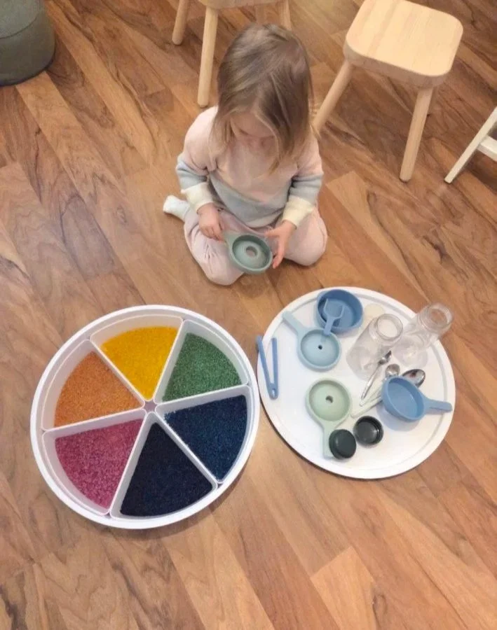 Ein kleines Mädchen sitzt auf dem Boden und spielt mit bunten Sandspielen und Wasser-Spiele, umgeben von verschiedenen Behältern und Werkzeugen auf zwei runden Tabletts.
