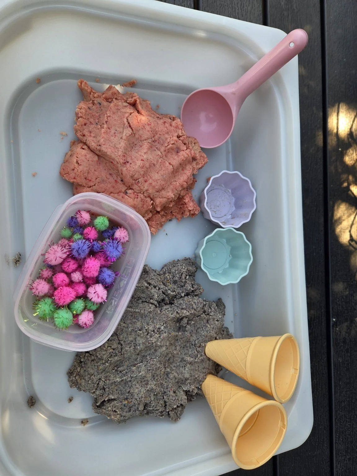 Kunstsand, Wassermelonen-Sandspielzeug, kleine Eistüte, bunte Pusteblumen, Steine und Kunststoffschalen auf einem Spieltisch.