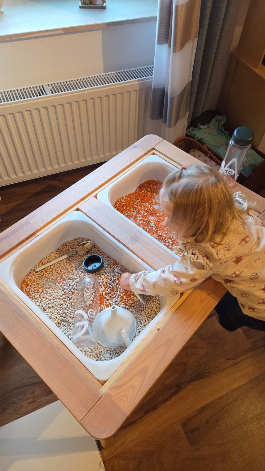 Ein Kind spielt an einem Tisch mit zwei Kinder-Sandkästen, die mit verschiedenen bunten getrockneten Hülsenfrüchten gefüllt sind.