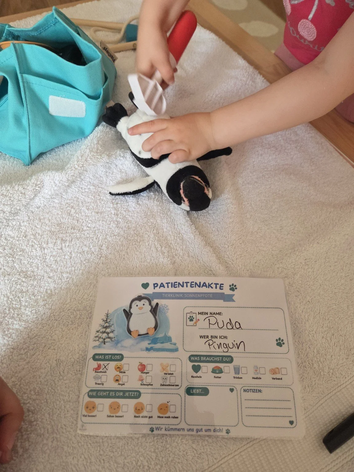 Ein kleines Kind legt eine Plüschpinguin auf eine weiße Decke. Neben dem Kind befindet sich eine blaue Tasche. Im Vordergrund liegt eine Patientenausfüllkarte mit einem Pinguin-Design, auf der handschriftlich 'Puda' als Name und 'Pinguin' als Tier eingetragen sind.