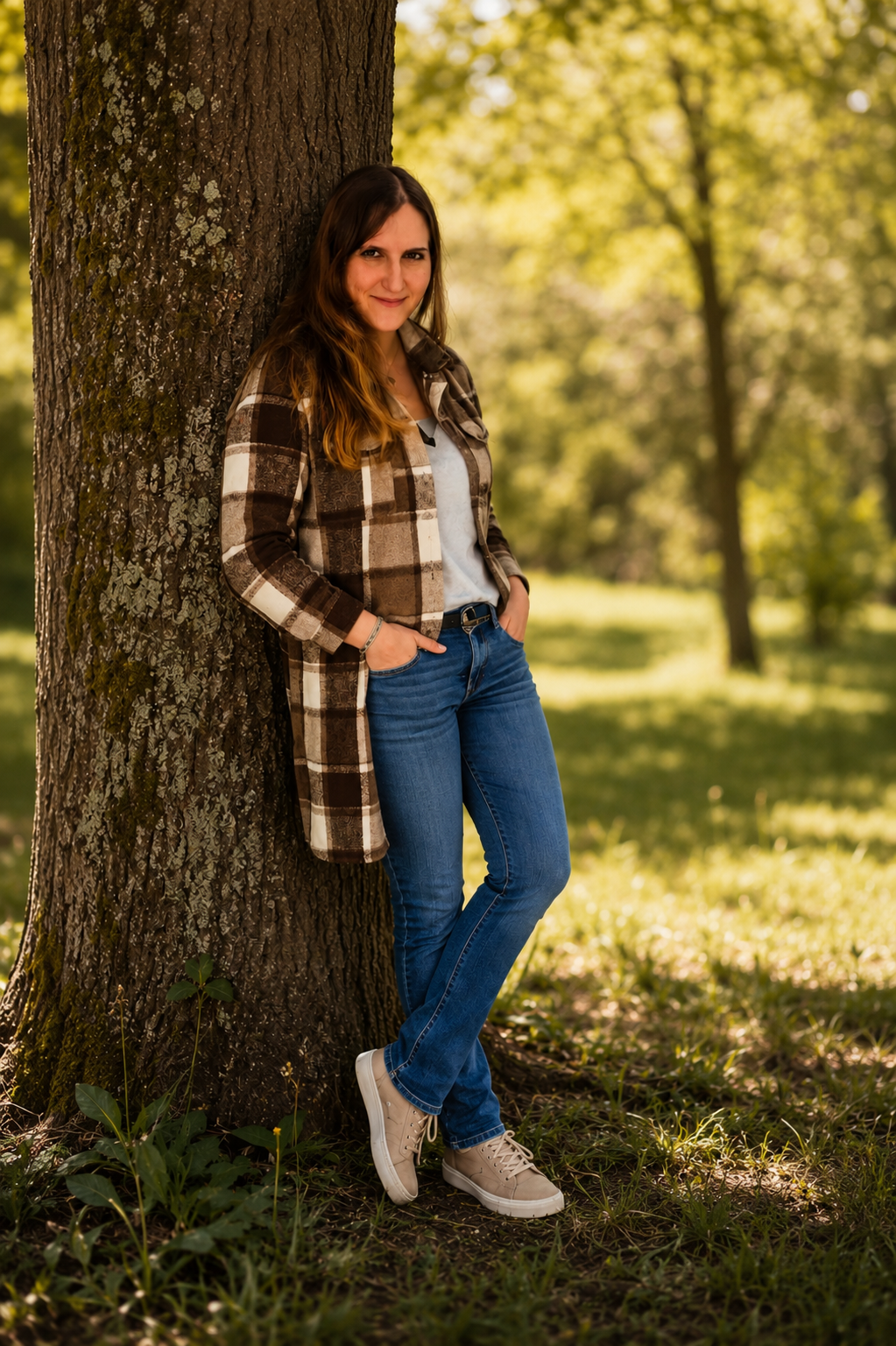 Junge Frau lehnt an einem Baum im Park, trägt grüne Jeans, ein graues Oberteil, eine braun-weiße kariert Jacke und beige Turnschuhe, umgeben von grüner Natur.