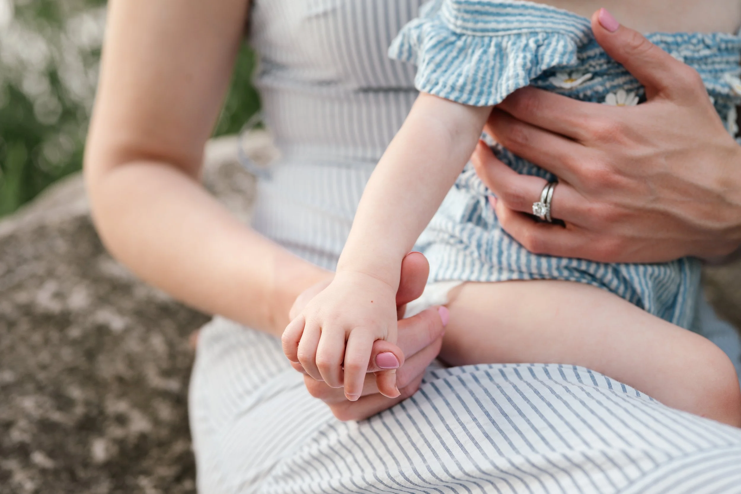 Close-up of an adult holding a child on their lap, with the child's arm and hand visible. The child's dress is blue and white striped with ruffles, and the adult is wearing a wedding ring. The background is blurred outdoor setting.