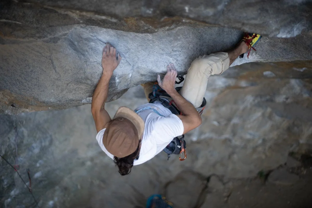 A person in white shirt and beige pants rock climbing on a vertical rock wall, wearing climbing gear including a harness and climbing shoes.