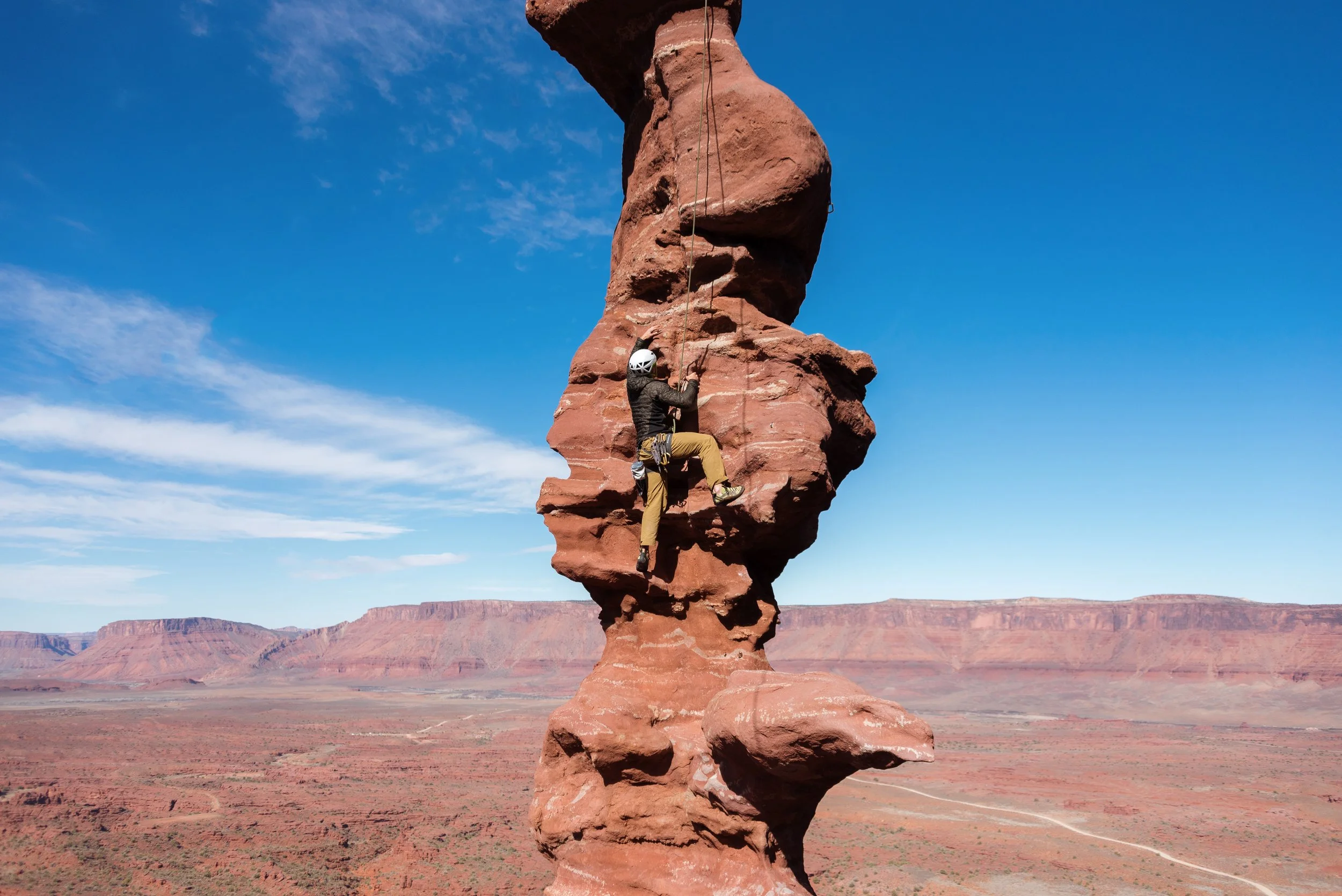 A person rock climbing on a tall, narrow, red sandstone formation in a desert landscape with canyon walls in the background under a blue sky.
