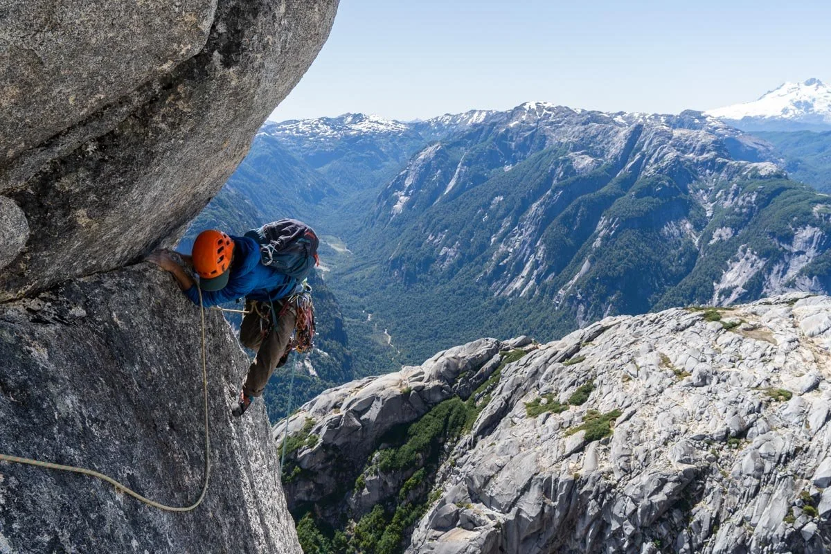 A rock climber ascending a steep cliff face with a scenic mountain valley and snow-capped peaks in the background.
